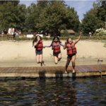 Three campers in red life jackets jumping into a lake
