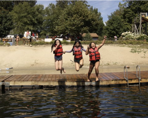 Three campers in red life jackets jumping into a lake