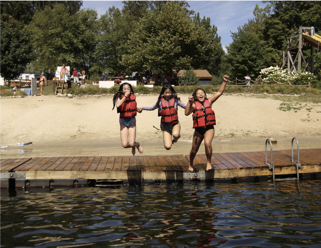 Three campers in red life jackets jumping into a lake