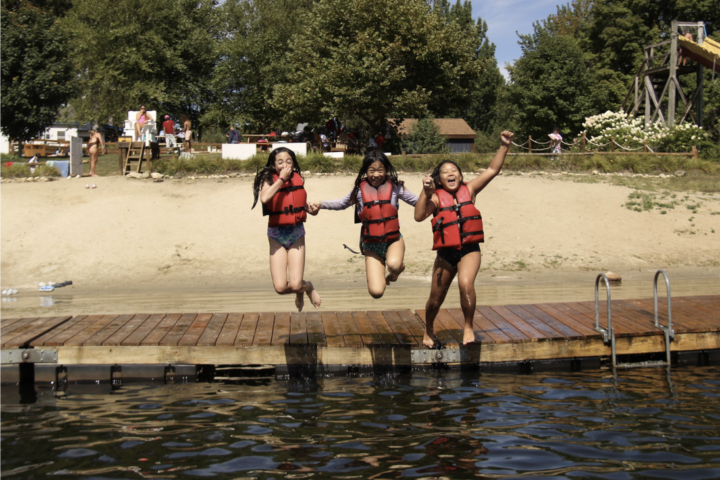 Three campers in red life jackets jumping into a lake